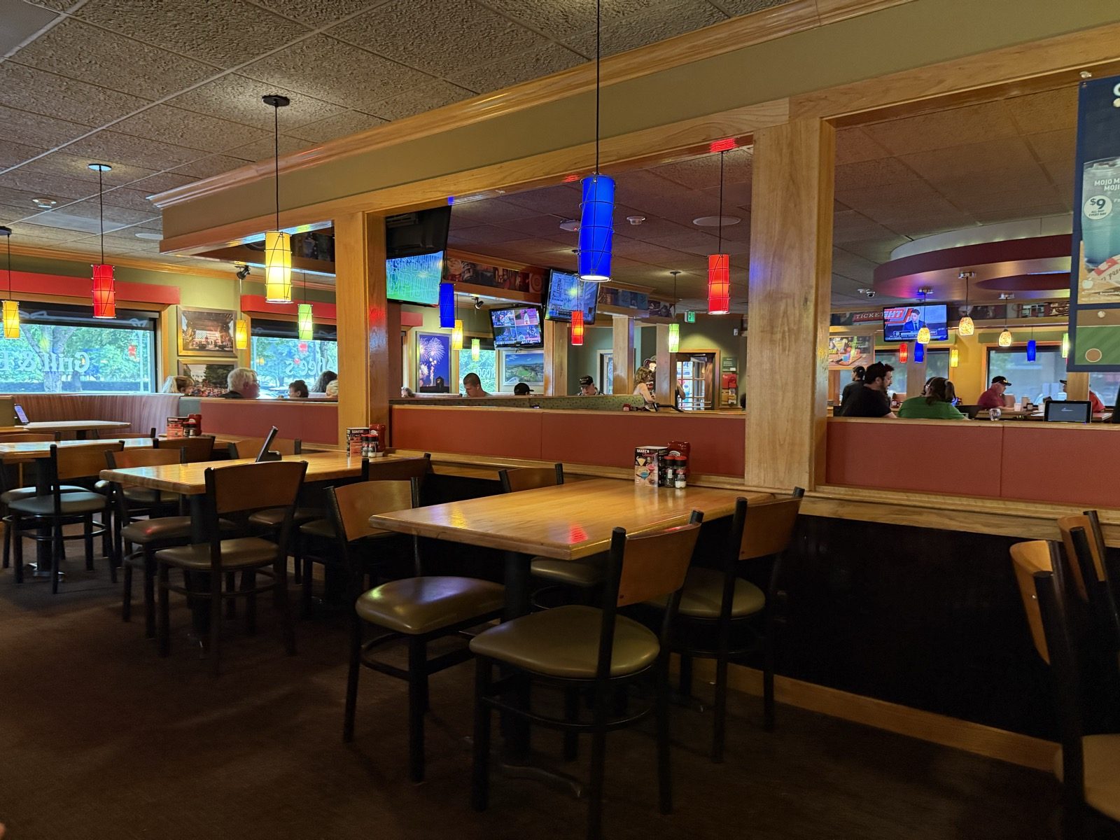 A brighter section of the dining room near the bar, hung with a row of colorful cylindrical paper lanterns in red, blue, and yellow, and several wall-mounted televisions, with diners seated at booths in the background