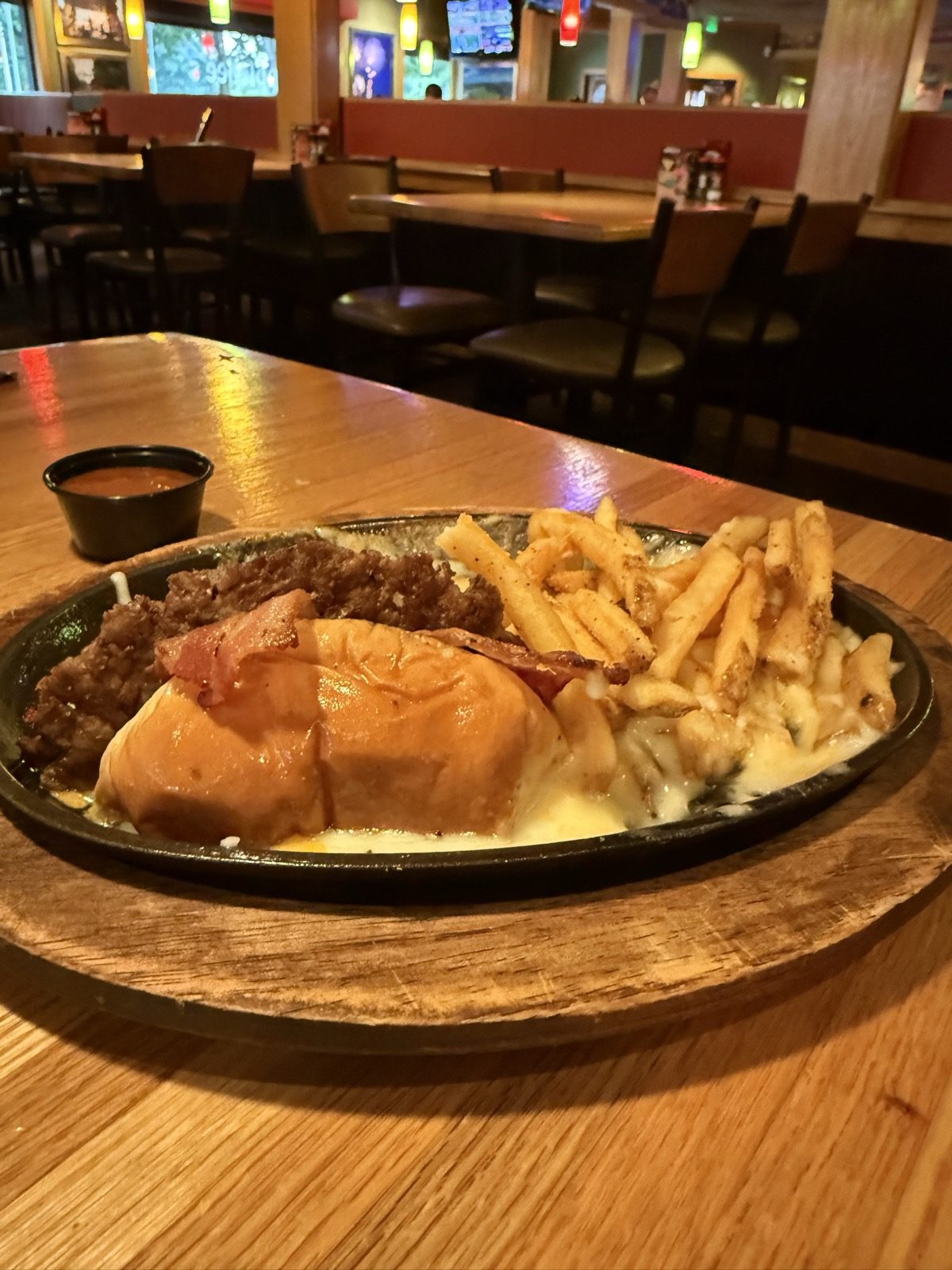 The O-M-Cheese Burger on a sizzling black cast-iron skillet — a brioche bun glistening with melted cheese sitting in a pool of molten queso, beside a pile of seasoned fries and a small cup of dipping sauce, on a wood table with the dining room softly lit behind it
