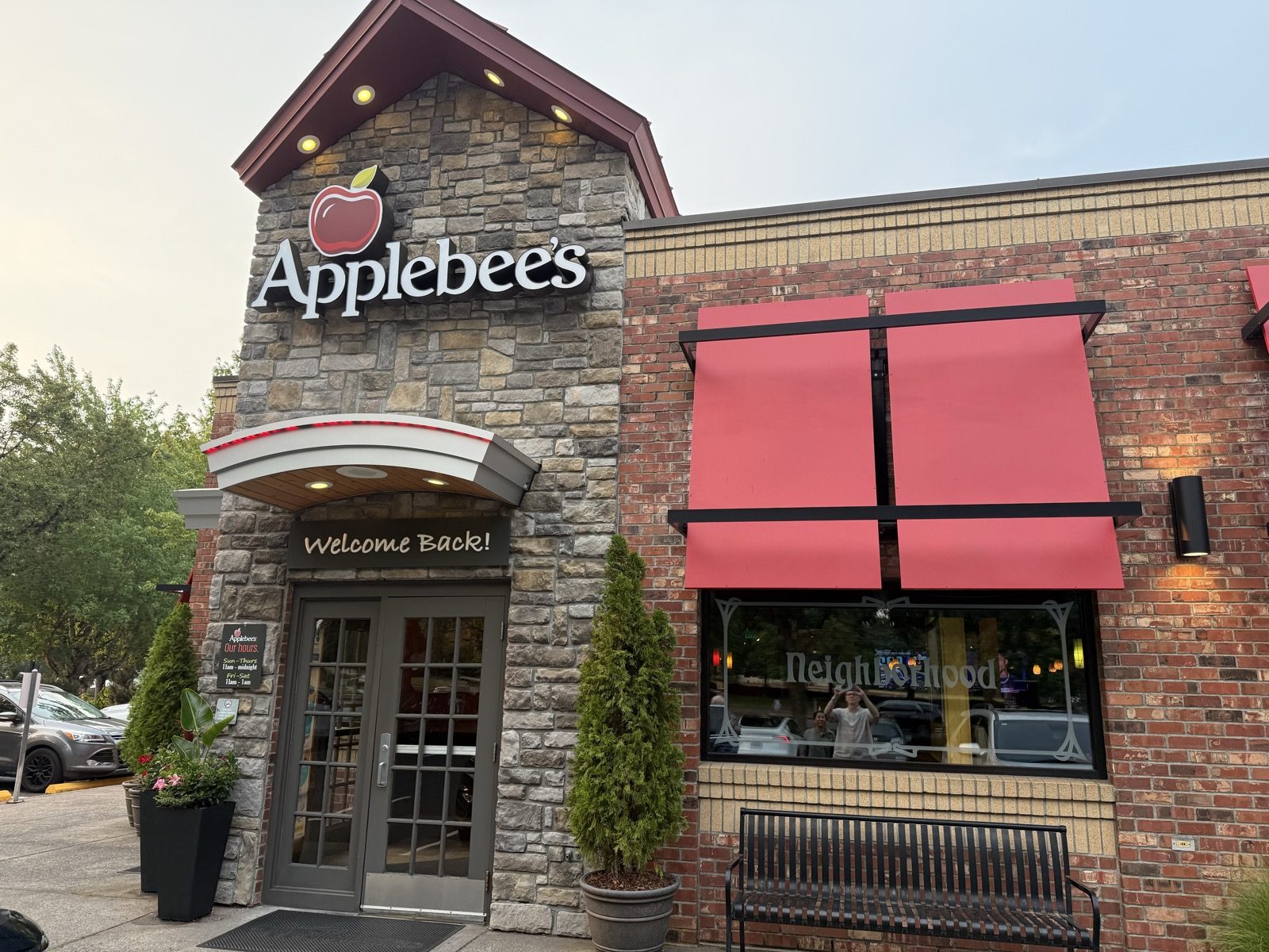 The Applebee's on SE 2nd Circle in Vancouver, Washington, photographed from the sidewalk at dusk — a stone-and-brick building with a peaked stone gable holding a lit red-and-white apple logo sign above glass doors under a curved awning reading "Welcome Back!", and beside it a window etched with the word "Neighborhood" next to a folded coral-pink awning