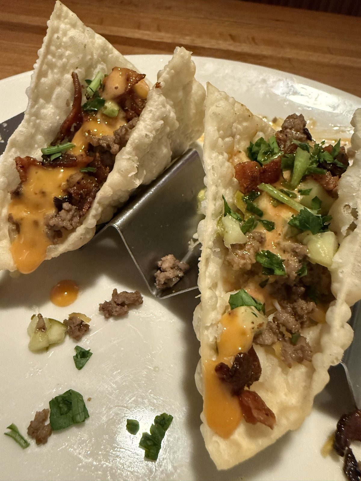 A white plate holding two Bacon Cheeseburger Wonton Tacos, crunchy wonton shells filled with seasoned ground beef, shredded cheese, bacon, and chopped pickle, drizzled with bright orange spicy honey mustard sauce, with crumbs on the plate where a third taco has already been eaten