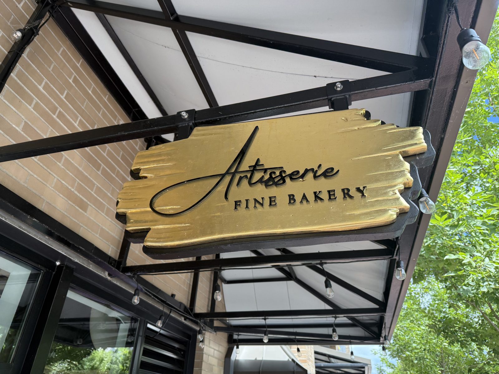 A gold hand-painted wooden sign reading Artisserie, Fine Bakery, hanging from black iron scrollwork under an awning