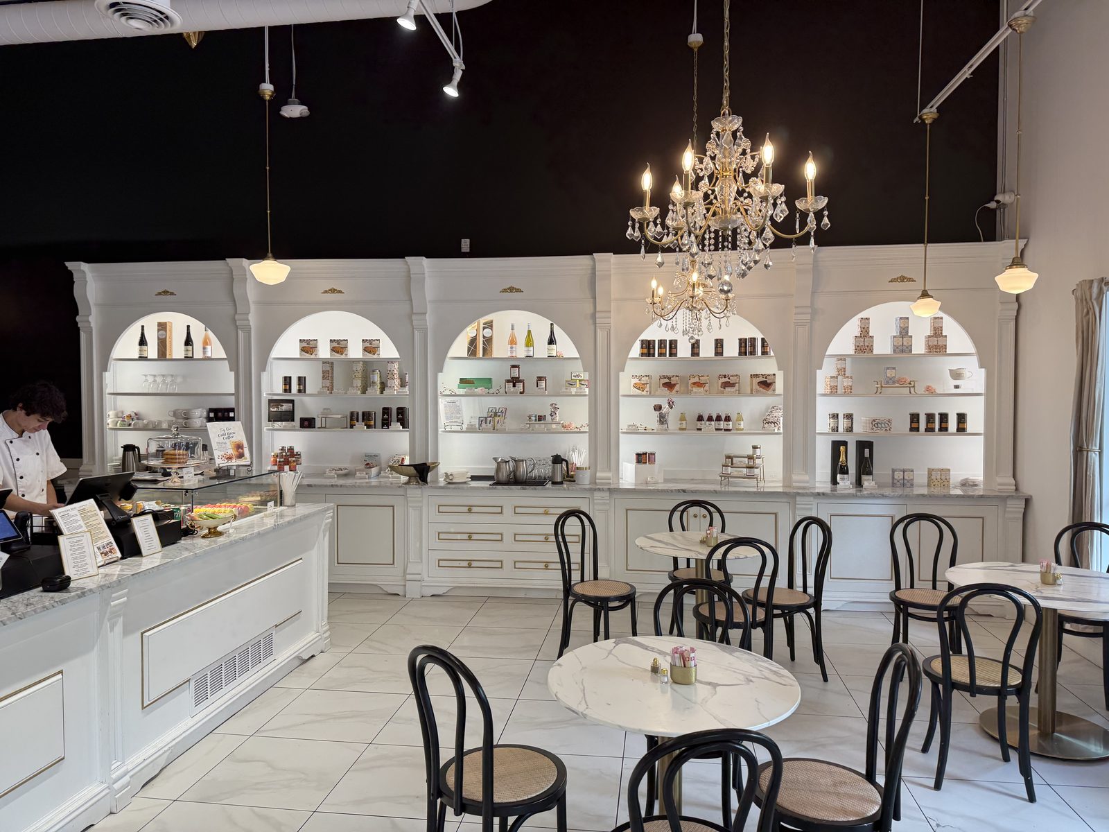 The white marble and gold-trimmed interior of Artisserie Fine Bakery, with an arched shelving display, a crystal chandelier, and small round marble bistro tables with black bentwood chairs