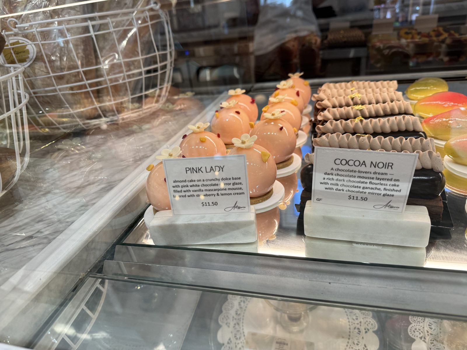 A pastry case with several domed Pink Lady desserts in pale pink mirror glaze, decorated with small white flowers, next to a card describing almond cake with vanilla mascarpone mousse and strawberry and lemon cream, priced at $11.50