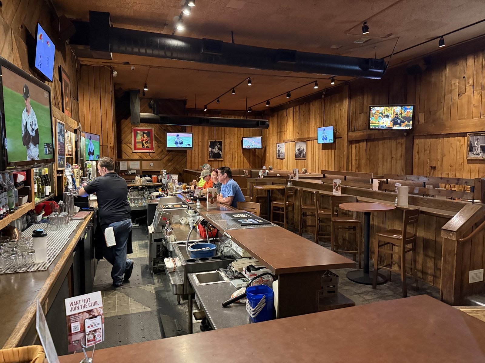 The wood-paneled bar and dining room at Black Angus Steakhouse, with a bartender in a black polo working behind the counter, several televisions mounted around the room showing a baseball game, three customers seated at the bar, and empty high-top tables in the foreground