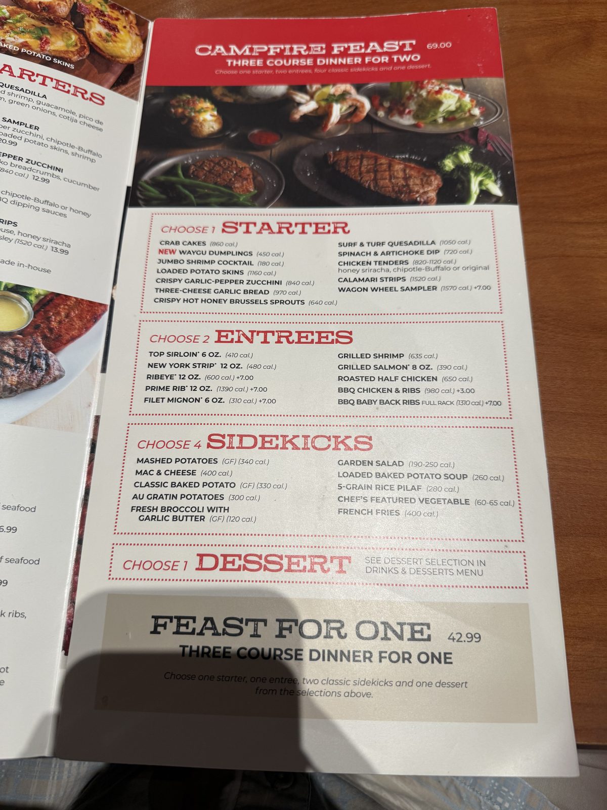 A red-bannered steakhouse menu page reading CAMPFIRE FEAST, THREE COURSE DINNER FOR TWO, $69.00, above a photograph of shared plates of steak, shrimp cocktail, salad and baked potato, followed by lists of starter, entrée and sidekick choices, with a Feast For One panel for $42.99 at the bottom