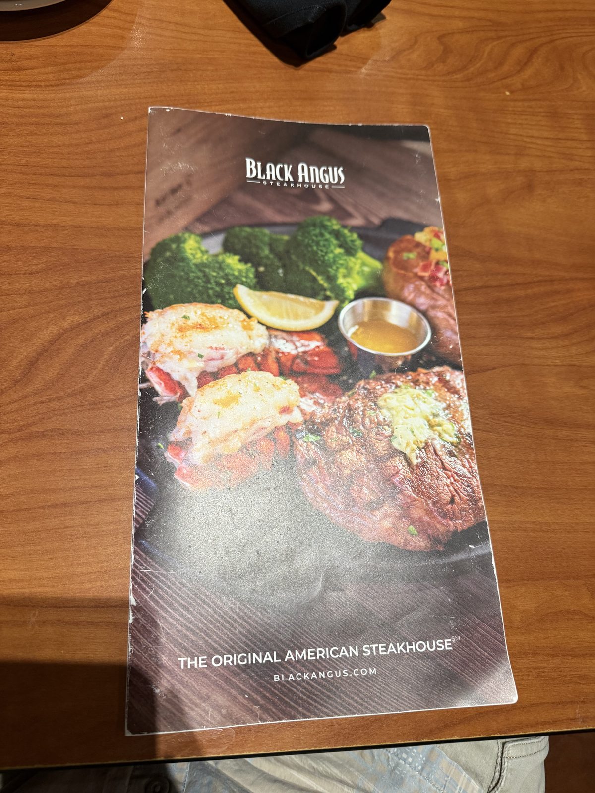 The Black Angus Steakhouse menu cover on a wood table, showing a plated ribeye and filet with garlic butter, two broiled lobster tails, broccoli and a lemon wedge, under the Black Angus Steakhouse title and the tagline The Original American Steakhouse