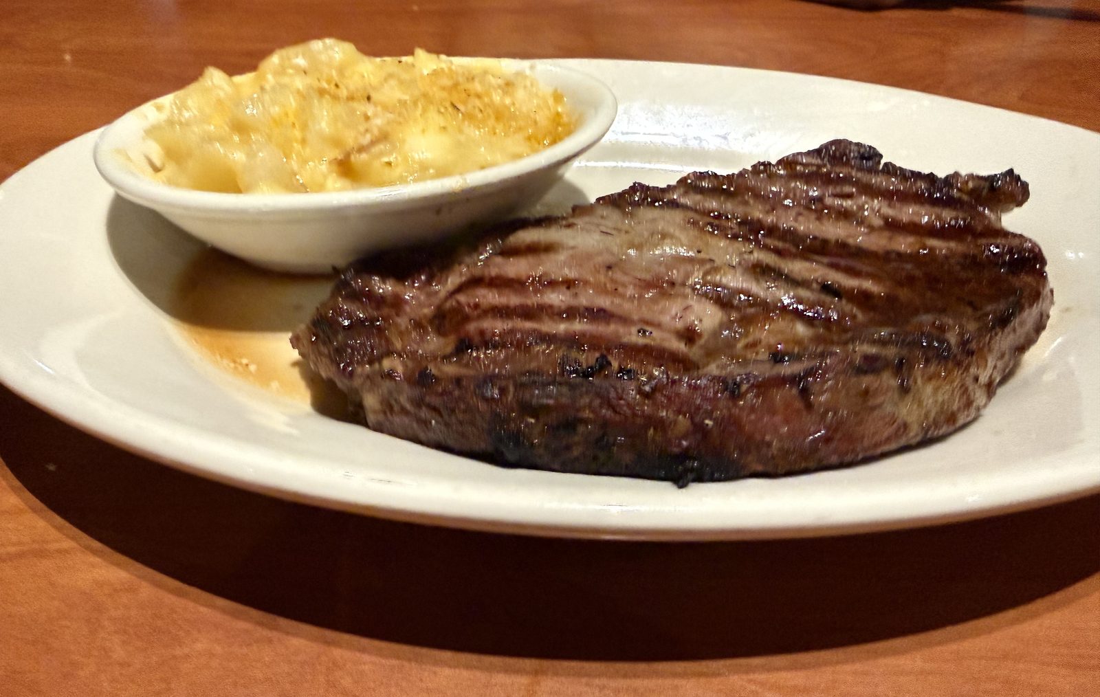 A thicker, evenly grilled New York strip steak on a white plate beside a bowl of au gratin potatoes