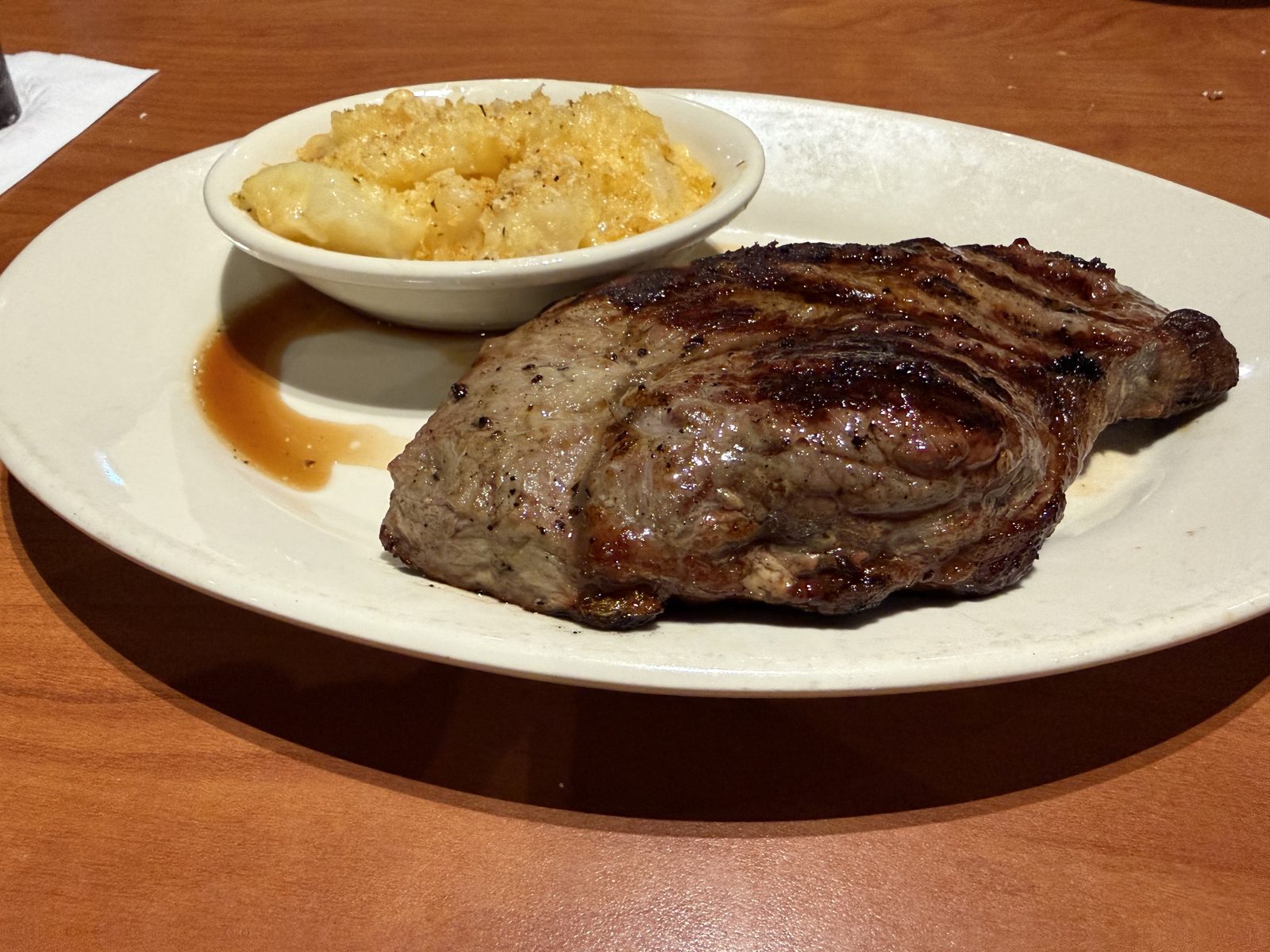 A thin, irregularly shaped grilled ribeye steak with dark char marks on a white oval plate, next to a bowl of golden au gratin potatoes, with a shallow pool of brown au jus underneath