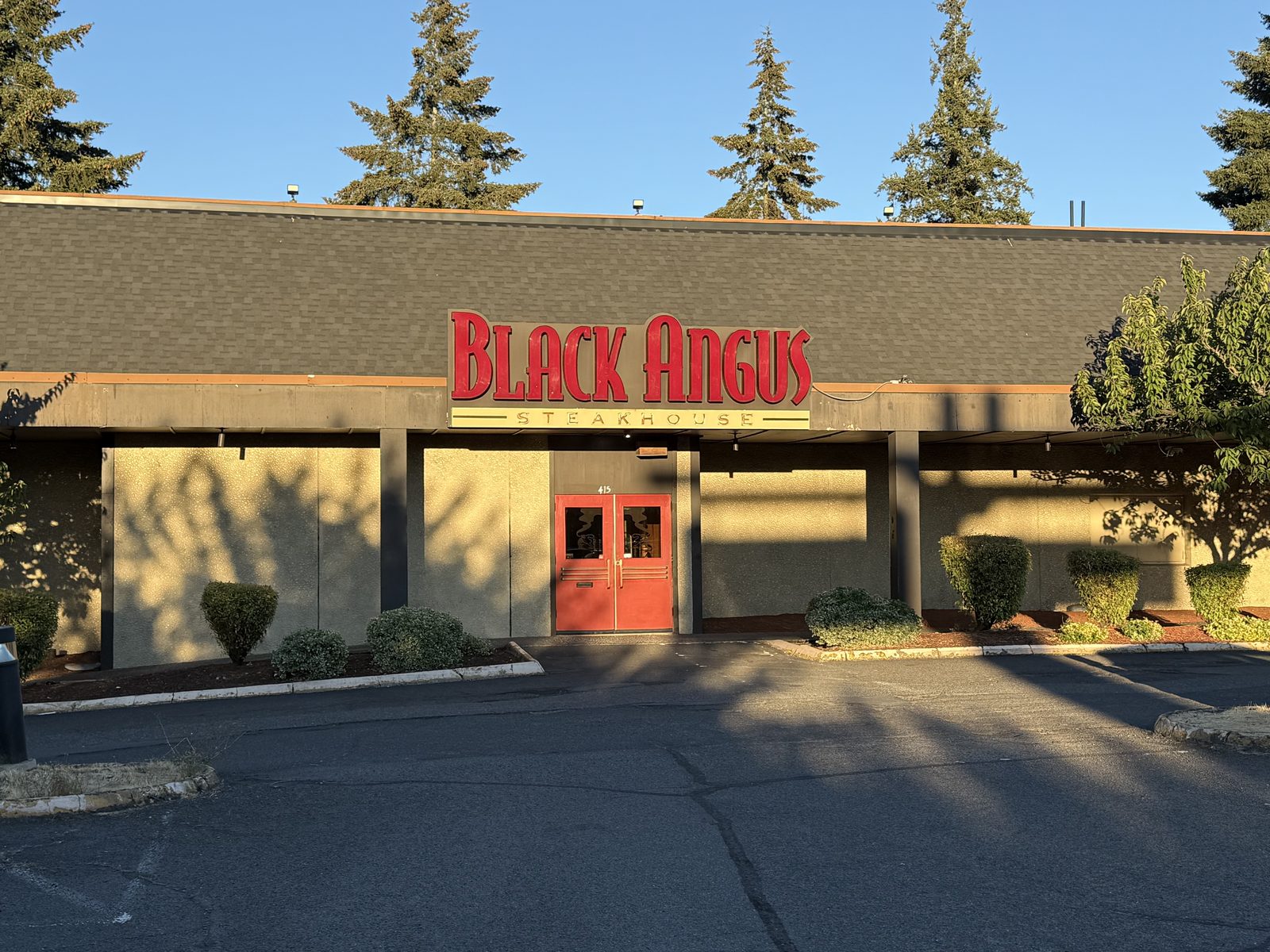 The Black Angus Steakhouse storefront on East 13th Street in Vancouver, Washington, photographed from the parking lot in the low gold light of early evening — a long tan building with a brown shingled mansard roof, a large red BLACK ANGUS STEAKHOUSE sign, red double doors marked 415 under the awning, and tall evergreen trees against a clear blue sky behind it