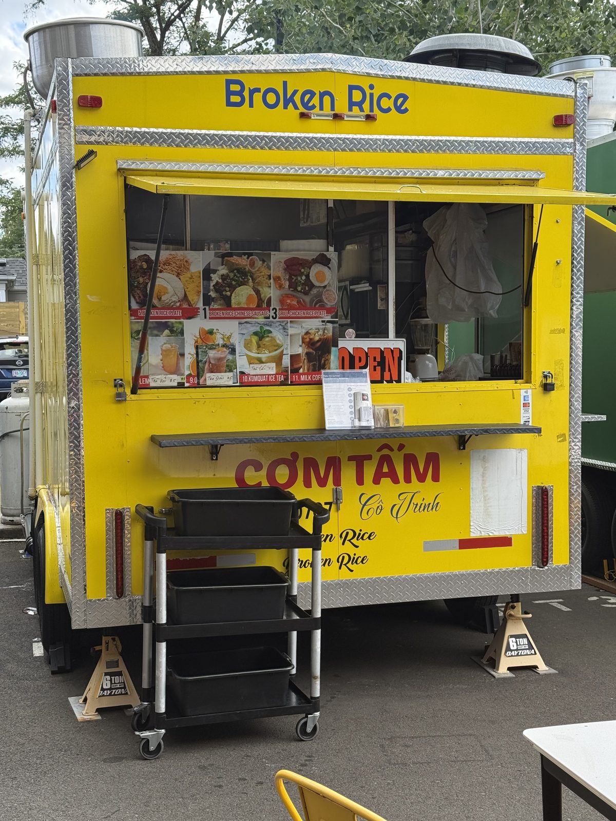 A yellow food cart trailer off 82nd Avenue in Portland, Oregon, with BROKEN RICE painted in blue across the top and COM TAM CO TRINH in red script below the service window, a photo menu board of rice plates and Vietnamese iced drinks taped inside, and a red OPEN sign lit in the window