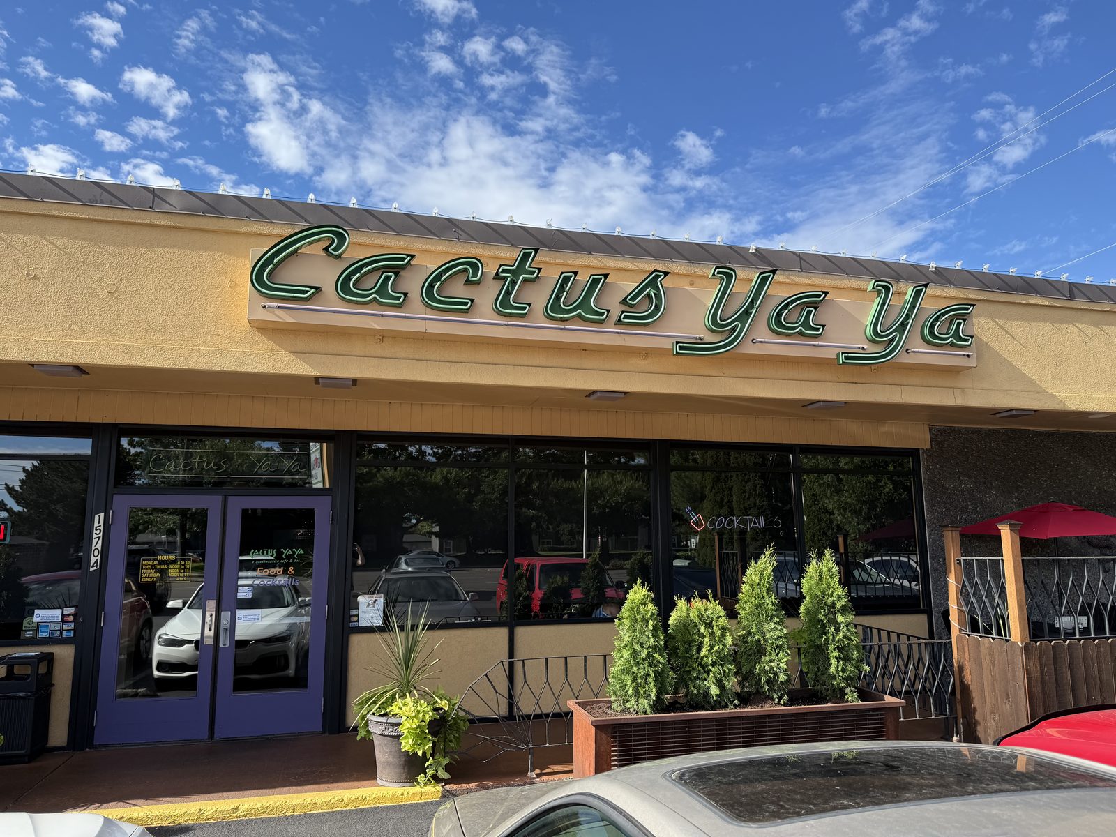 The green neon script sign reading Cactus Ya Ya above the entrance of a stucco building, with potted junipers and a red umbrella on the patio out front