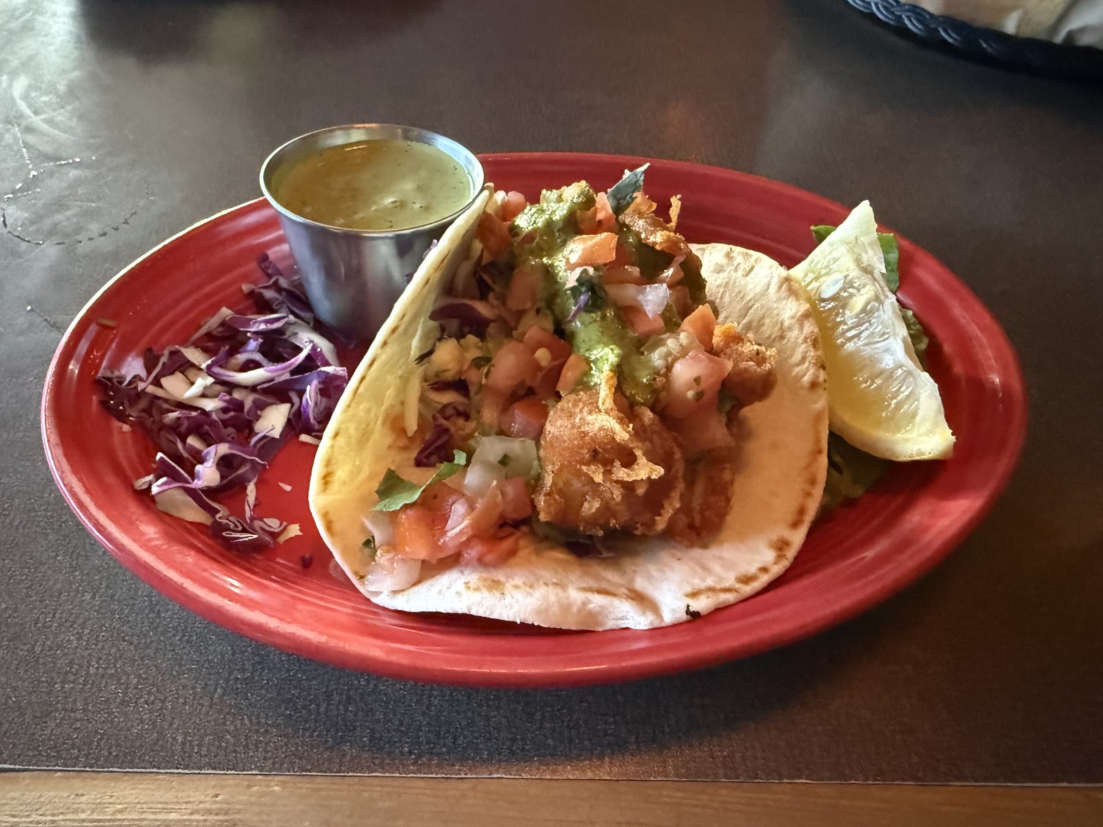 A fried fish taco on a red plate, topped with purple cabbage slaw, pico de gallo and avocado crema, with a lime wedge and a small metal cup of green sauce