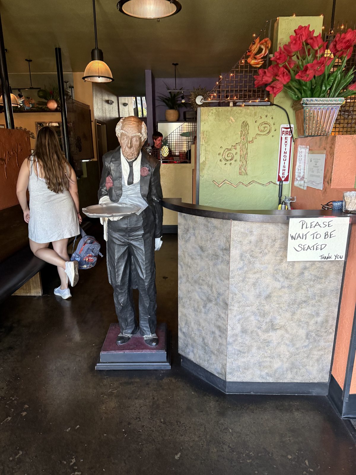 The host stand at Cactus Ya Ya, with a life-size statue of a waiter in a white coat holding a serving tray, next to a hand-lettered please-wait-to-be-seated sign and a vase of red flowers