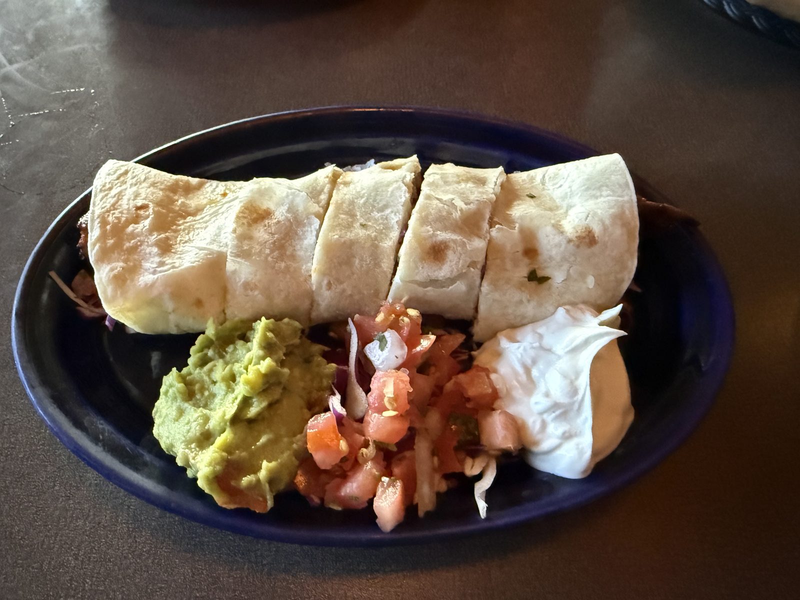 A grilled steak yaya roll sliced into pieces on a dark blue plate, steak and melted cheese visible at the folds, served with guacamole, pico de gallo and sour cream