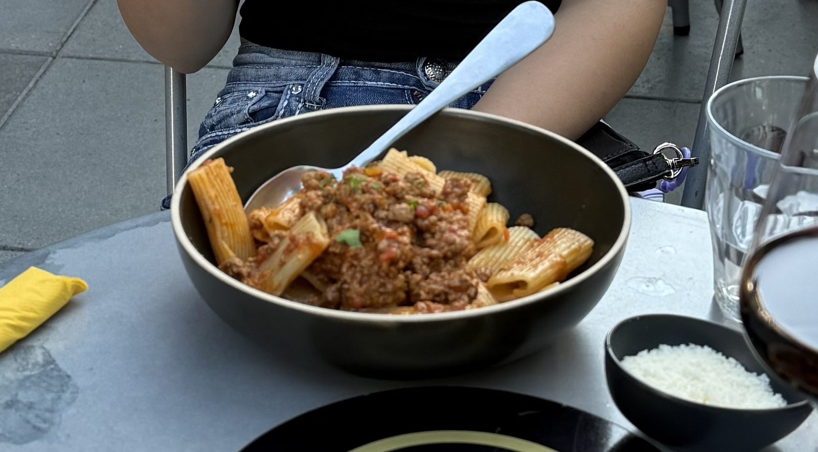 A dark olive bowl of rigatoni Bolognese at Chuckie Pies, ridged tubes of pasta under a coarse ground meat ragu flecked with green parsley, a serving spoon resting in the bowl and a small black dish of grated parmesan beside it