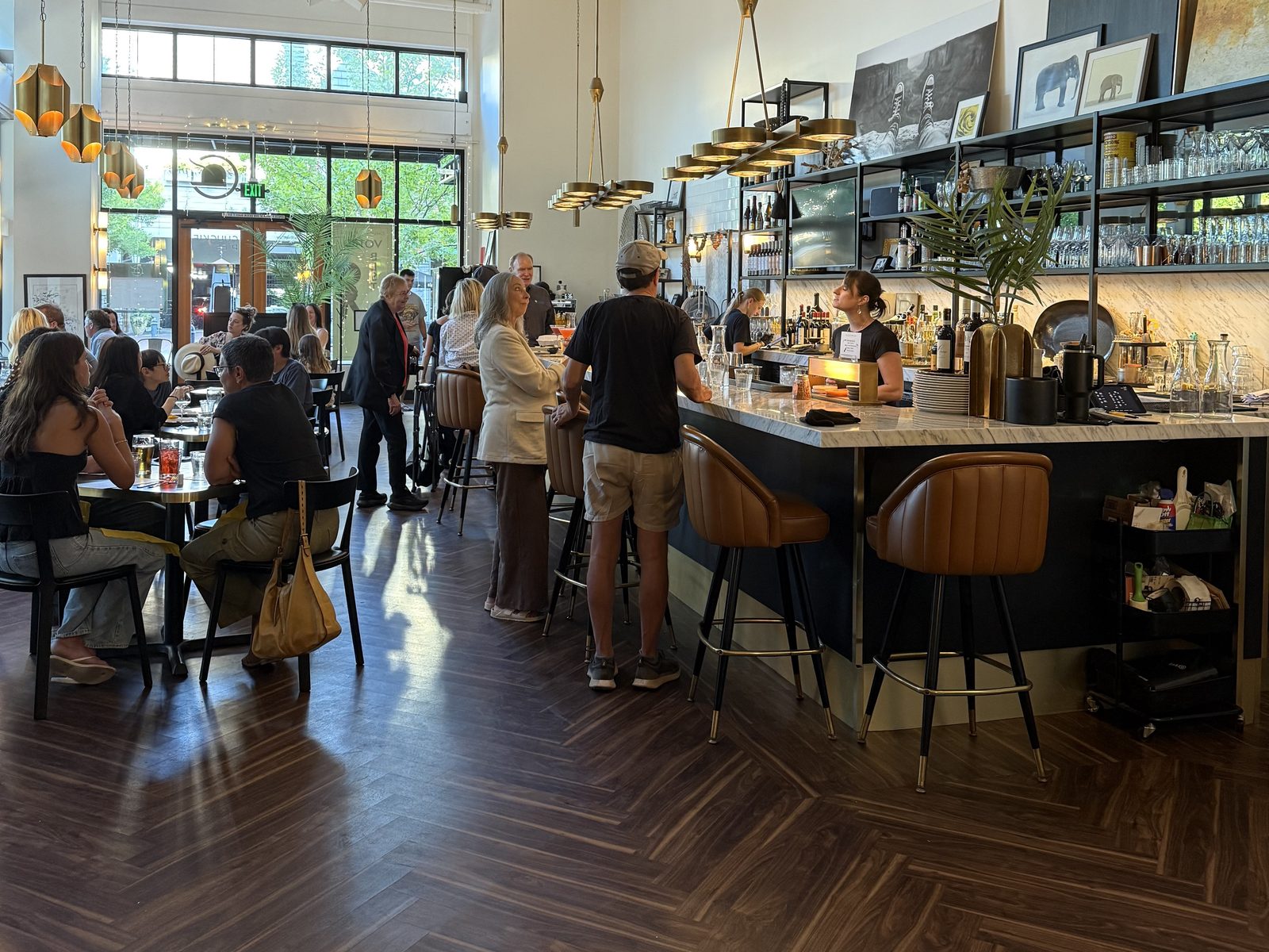The dining room at Chuckie Pies on a busy Thursday evening, dark walnut herringbone floor, double-height white ceiling with exposed ductwork, brass pendant lights and a marble-topped bar with tan leather stools, all of them occupied, and floor-to-ceiling windows onto First Street