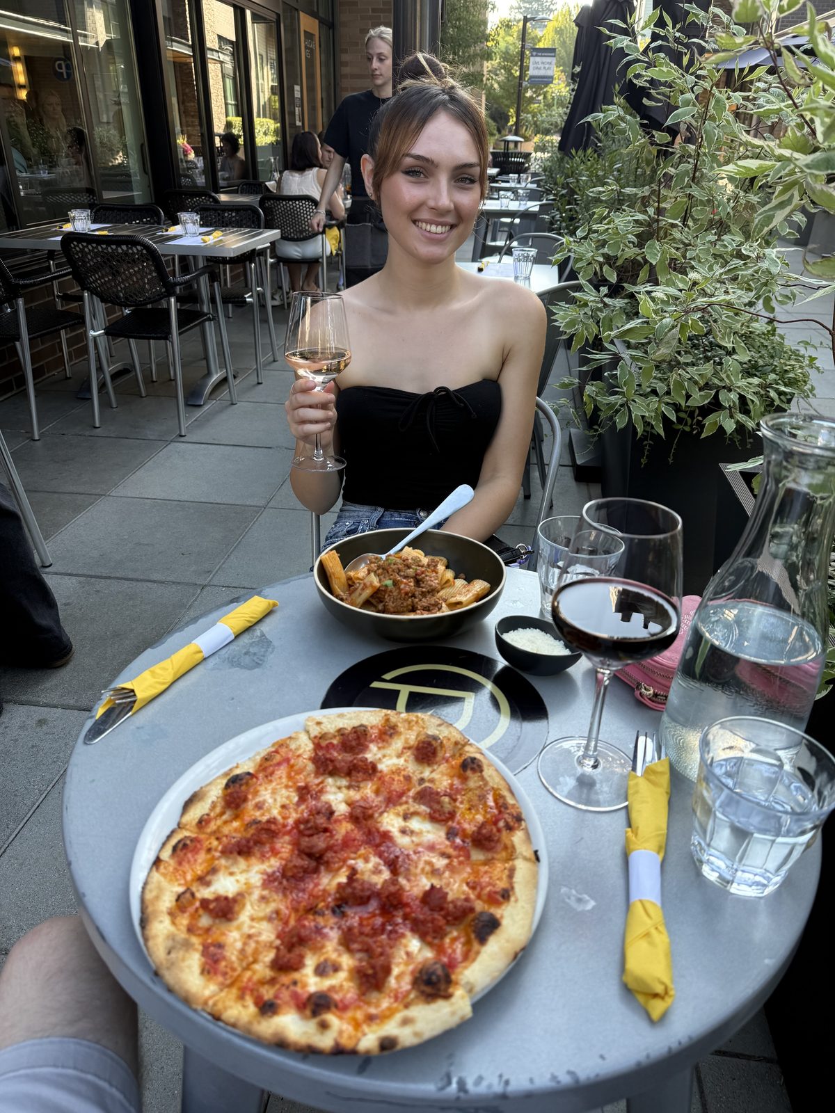 Dinner on the patio at Chuckie Pies in Lake Oswego, Oregon: a young woman sitting across a round grey table raising a glass of wine, a dark bowl of rigatoni Bolognese in front of her, and a ten-inch Neapolitan sausage pizza on a white plate in the foreground beside a carafe of water and two yellow rolled napkins