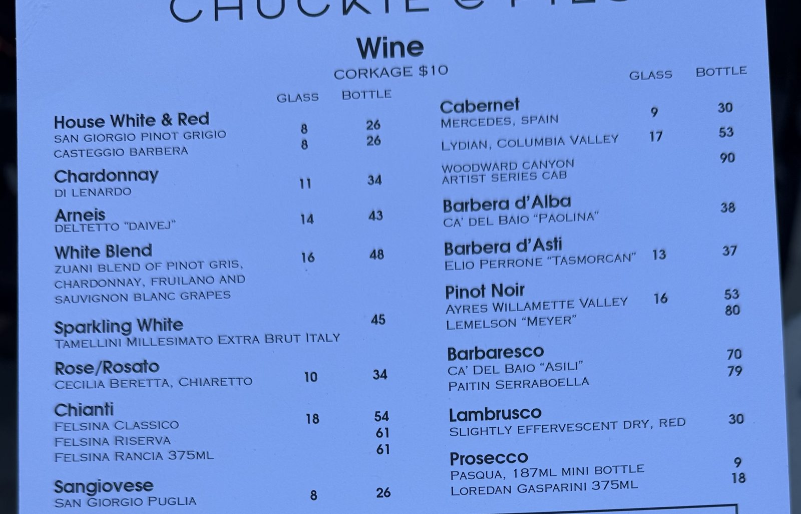 The wine section of the Chuckie Pies drinks menu on pale blue paper, listing house white and red, Chardonnay, Arneis, a white blend, sparkling, rosato, three Felsina Chiantis, Sangiovese, Cabernet, Barbera d'Alba, Barbera d'Asti, Pinot Noir, Barbaresco, Lambrusco and Prosecco, above a boxed split size wine bottle for 2 section
