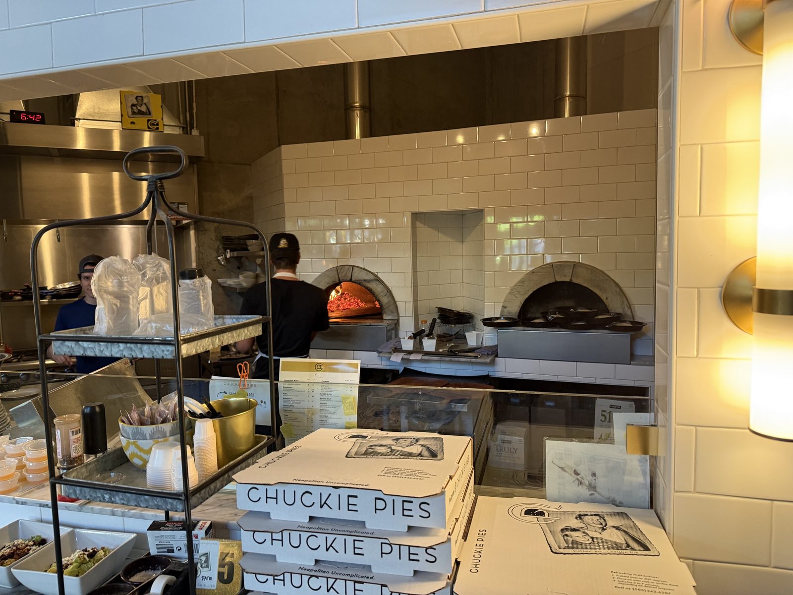 The open kitchen at Chuckie Pies, two arched wood-fired ovens set into a cream tiled wall with a fire burning in the left one, a cook working at the pass, and a stack of pizza boxes printed Chuckie Pies, Neapolitan Uncomplicated, with a vintage photograph captioned Esther and Chuckie Pie