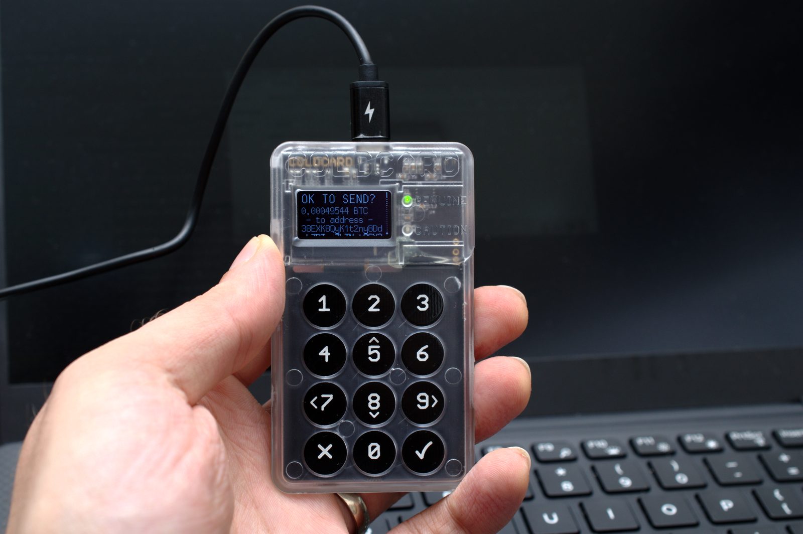 A hand holding a Coldcard hardware wallet, its screen reading OK TO SEND with a bitcoin amount and address, its numeric keypad below