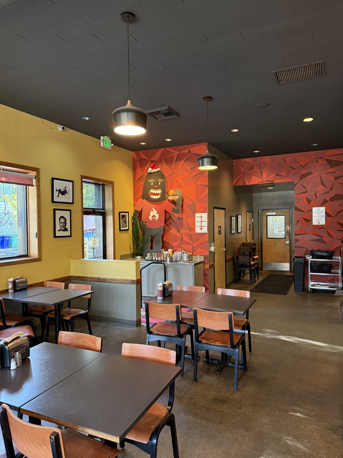 The dining room at Dick's Primal Kitchen — mustard-yellow and terracotta walls, a mural of a yeti in a red T-shirt holding a burger, dark two-top tables with wood-and-steel chairs, a self-serve water and cutlery station, and framed black-and-white portraits