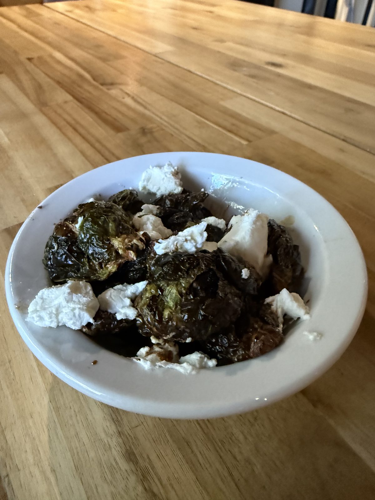 A white bowl of roasted Brussels sprouts, dark and blistered at the edges, with balsamic reduction pooled underneath and chunks of white goat cheese scattered over the top