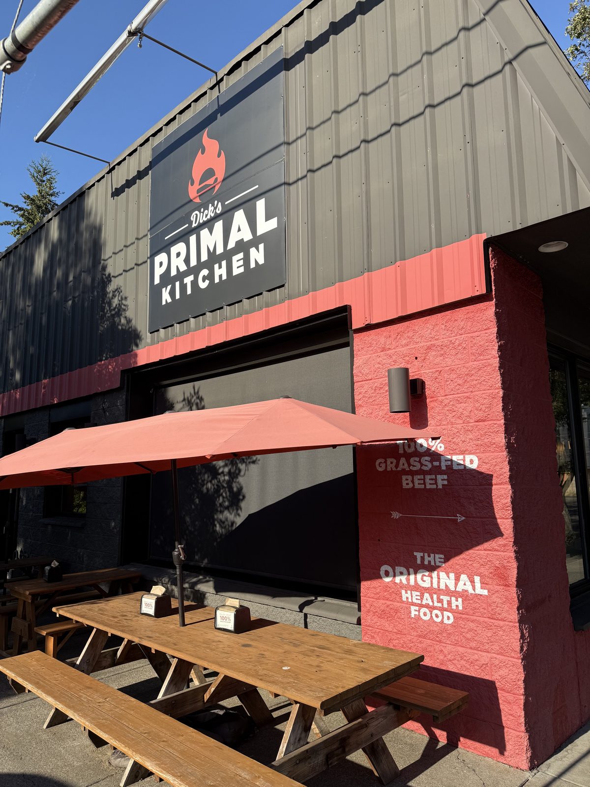 The Dick's Primal Kitchen storefront on SE Woodstock Blvd — a dark corrugated-metal facade with a black sign showing a red flame-and-burger logo, a terracotta-red painted wall reading 100% GRASS-FED BEEF and THE ORIGINAL HEALTH FOOD, and long wooden picnic tables under a red umbrella out front