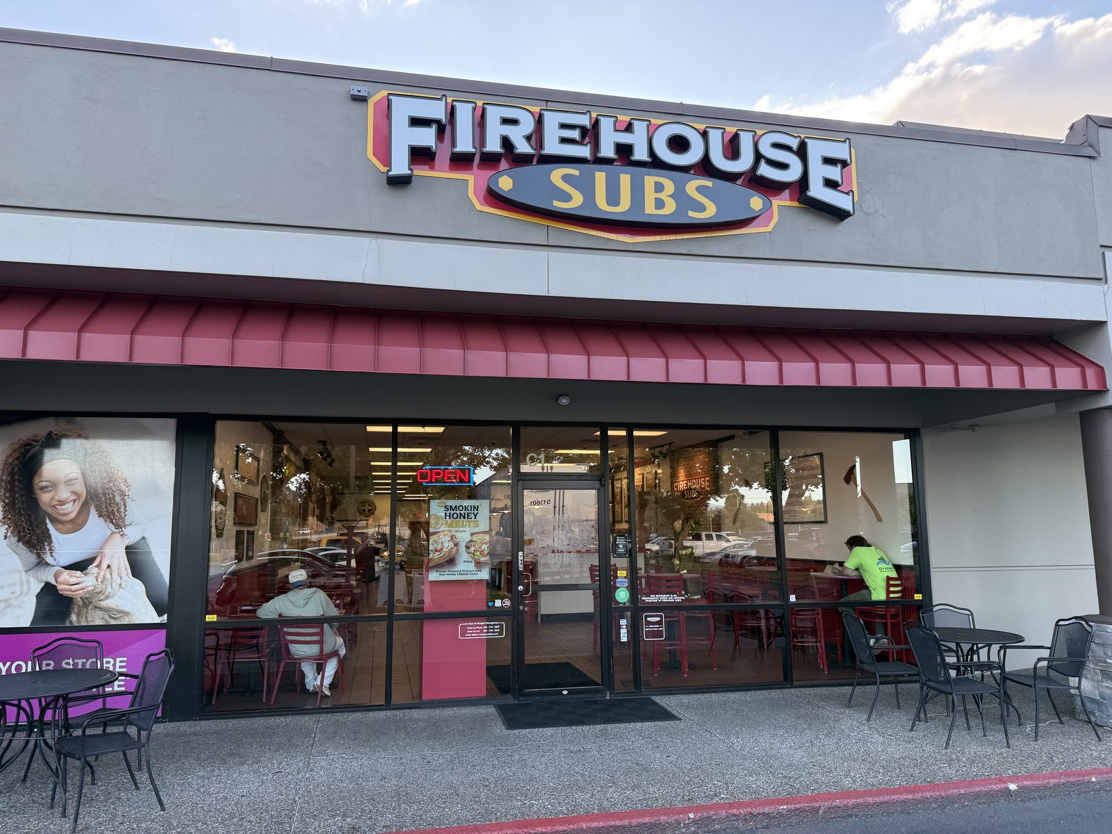 The Firehouse Subs storefront at dusk, red block letters on a beige facade above a red awning, an OPEN neon sign glowing in the window and a few customers seated inside