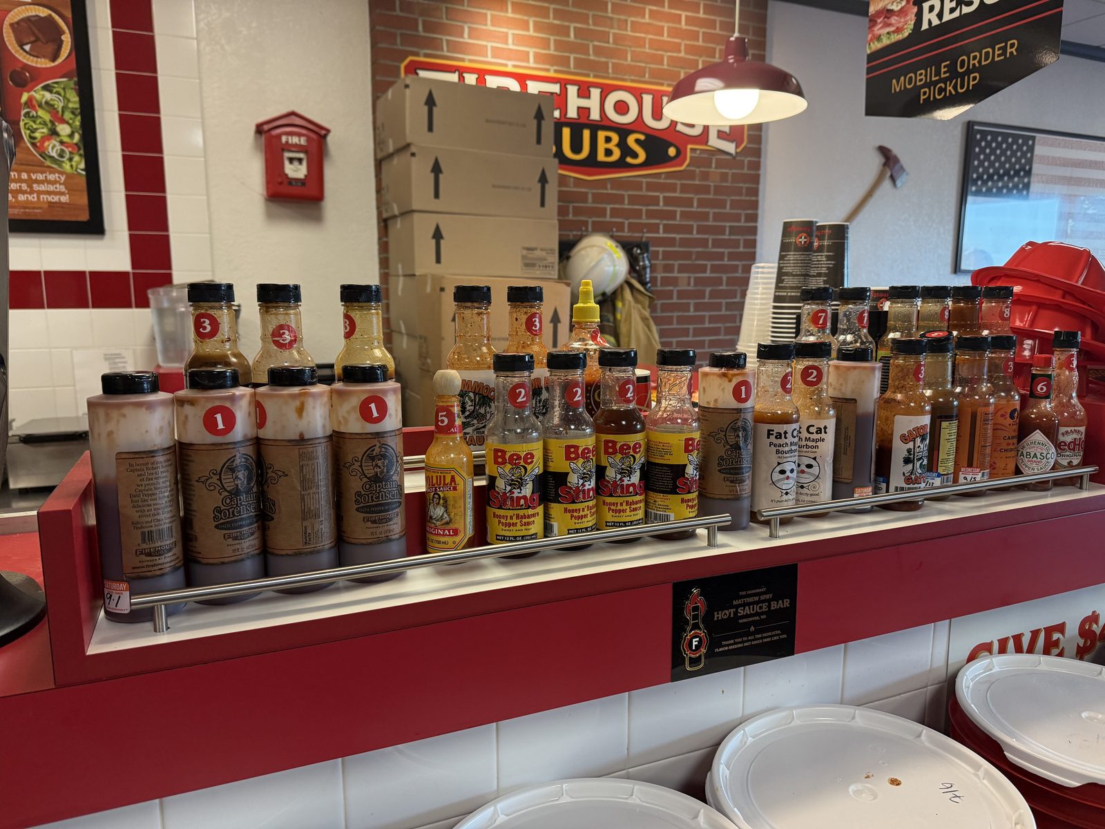 The hot sauce bar at Firehouse Subs, a chrome rail holding numbered bottles including several jugs of Captain Sorensen's sauce at heat level 1, beneath a plaque reading Hot Sauce Bar