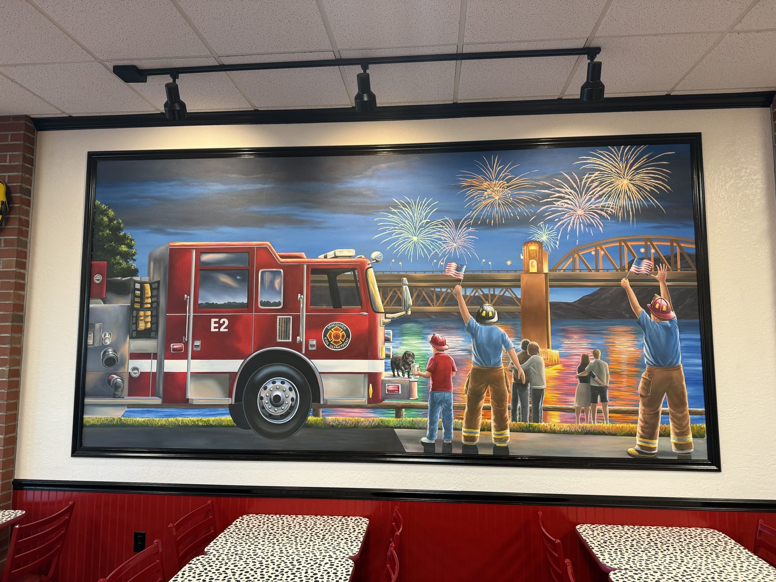 A large painted mural inside Firehouse Subs of a red fire engine numbered E2 parked beside a river at night, with two firefighters and a small dog watching fireworks burst over a bridge in the background