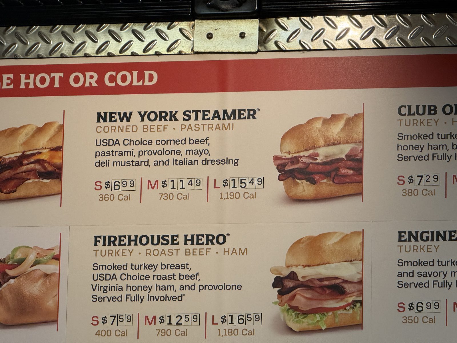 A close-up of the Firehouse Subs menu board listing the New York Steamer — corned beef and pastrami, USDA Choice corned beef, pastrami, provolone, mayo, deli mustard and Italian dressing — priced small $6.99, medium $11.49, large $15.49