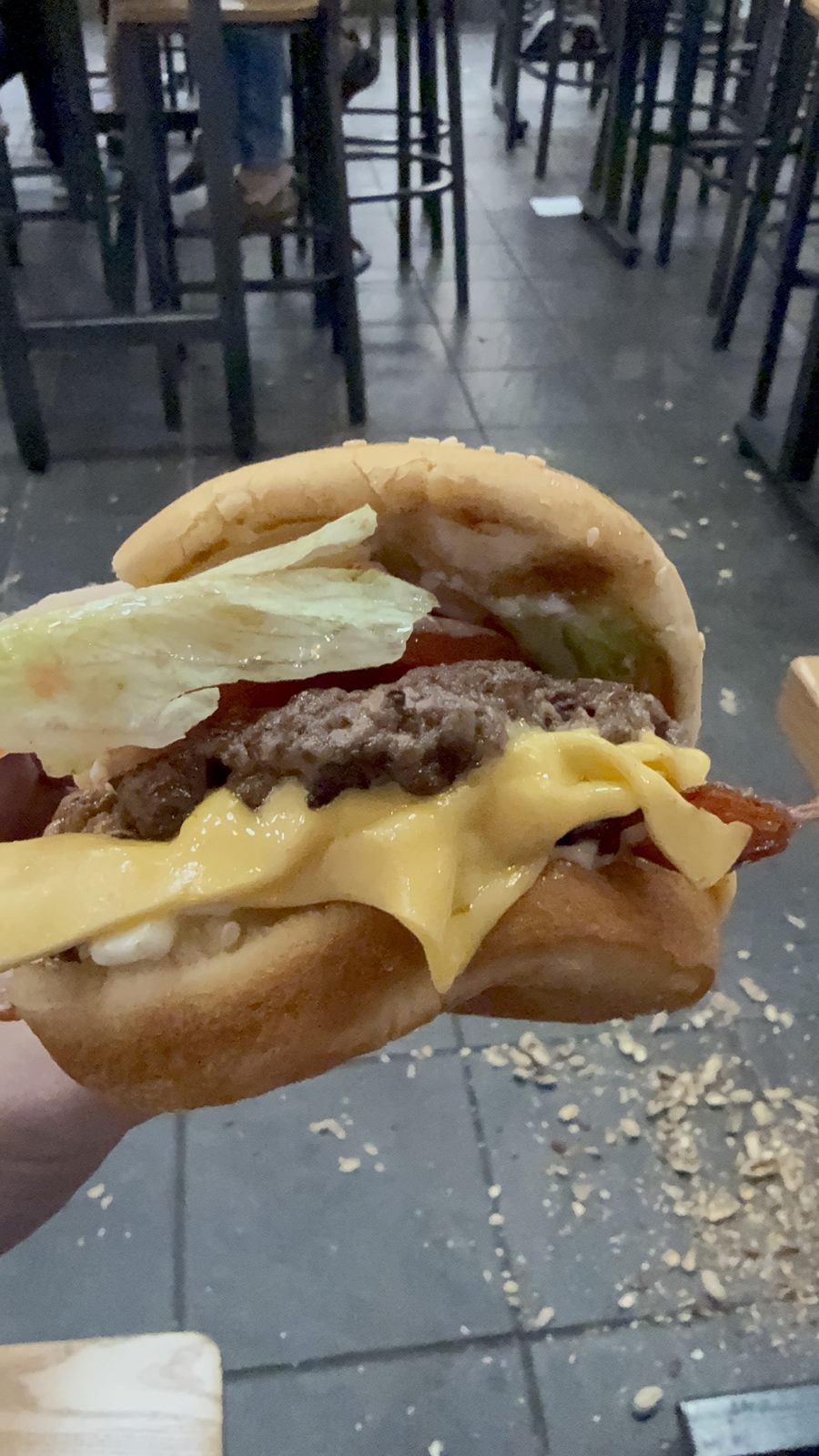 A close-up of a bacon cheeseburger from Five Guys held up for the camera, showing a beef patty, melted American cheese, a strip of bacon, lettuce and tomato between a bun, with peanut shells scattered on the floor below