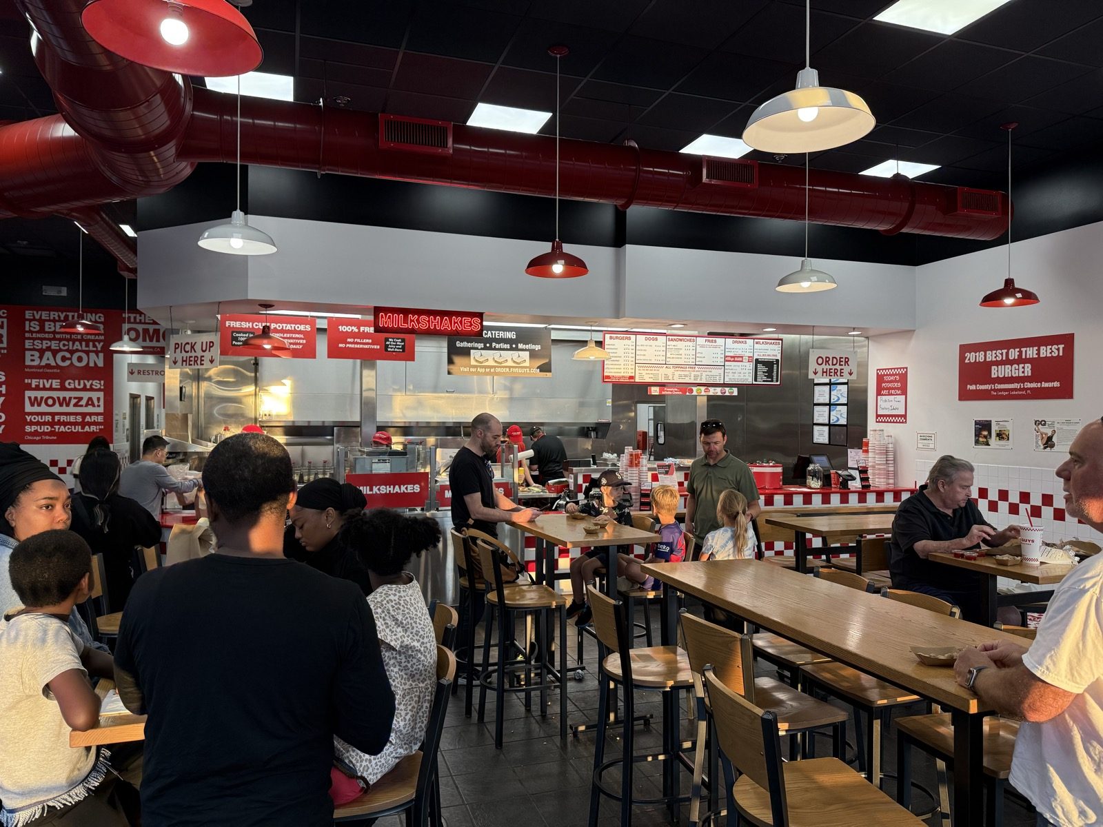 A crowded Five Guys dining room in Vancouver, Washington at evening, with families seated at wooden tables and a line of customers waiting near the counter, red-and-white checkered tile and a '2018 Best of the Best Burger' award sign on the wall