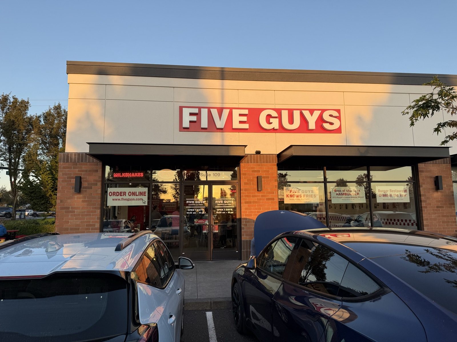 The lit red-and-white Five Guys sign above the entrance of a Vancouver, Washington location at golden hour, with two cars parked in front and a warm evening sky overhead