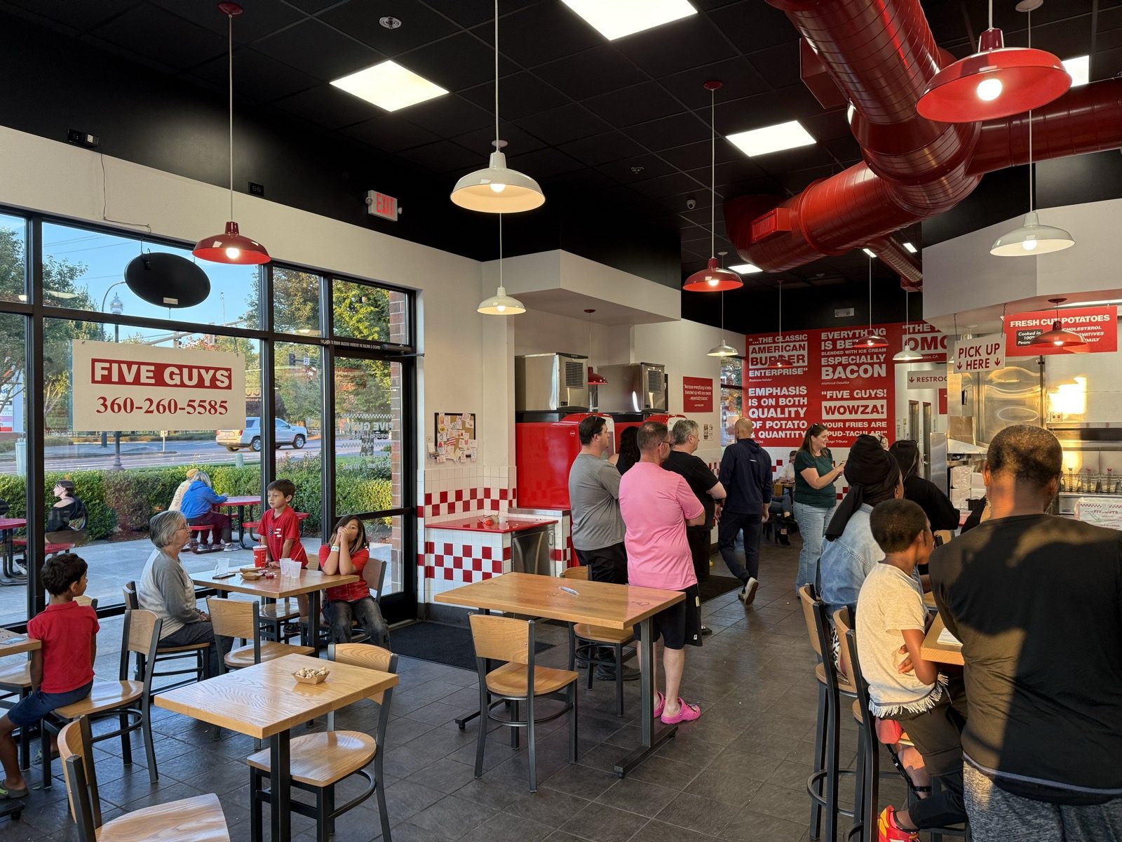 The window of a Five Guys restaurant in Vancouver, Washington printed with the store name and phone number, with customers waiting in line and seated at tables inside during a busy evening
