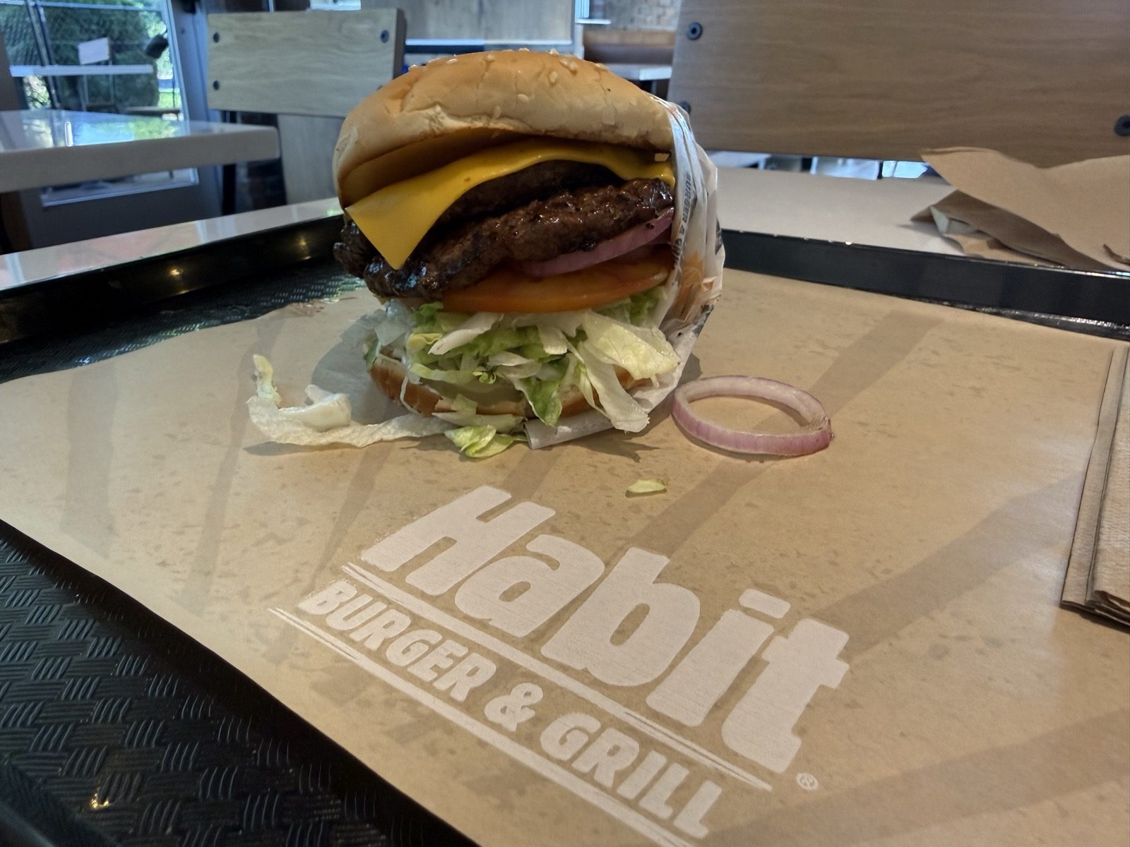 A Double Char cheeseburger from Habit Burger and Grill sitting on a paper wrapper printed with the Habit logo, showing two chargrilled beef patties, melted cheese, tomato and shredded lettuce, with a ring of red onion set aside next to it