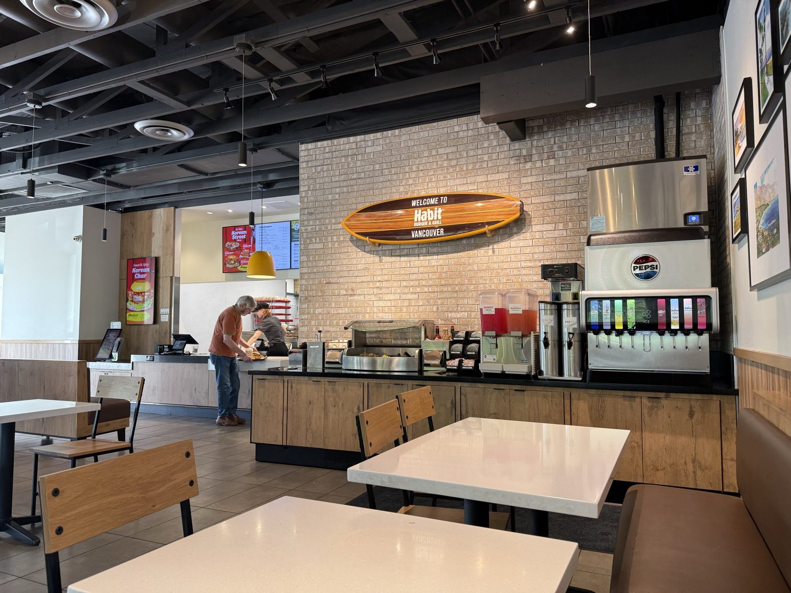 The counter area inside Habit Burger and Grill in Vancouver, Washington, with a wooden surfboard mounted on a brick wall reading Welcome to Habit Burger and Grill Vancouver, above a drink station and ice machine