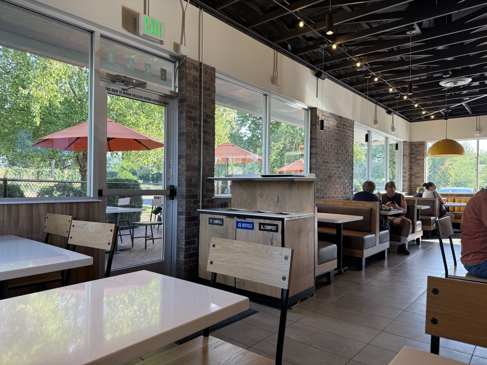 The bright dining room at Habit Burger and Grill in Vancouver, Washington, with white tables, wood-backed chairs, exposed dark ceiling beams and string lights, a glass door open to a patio with orange umbrellas