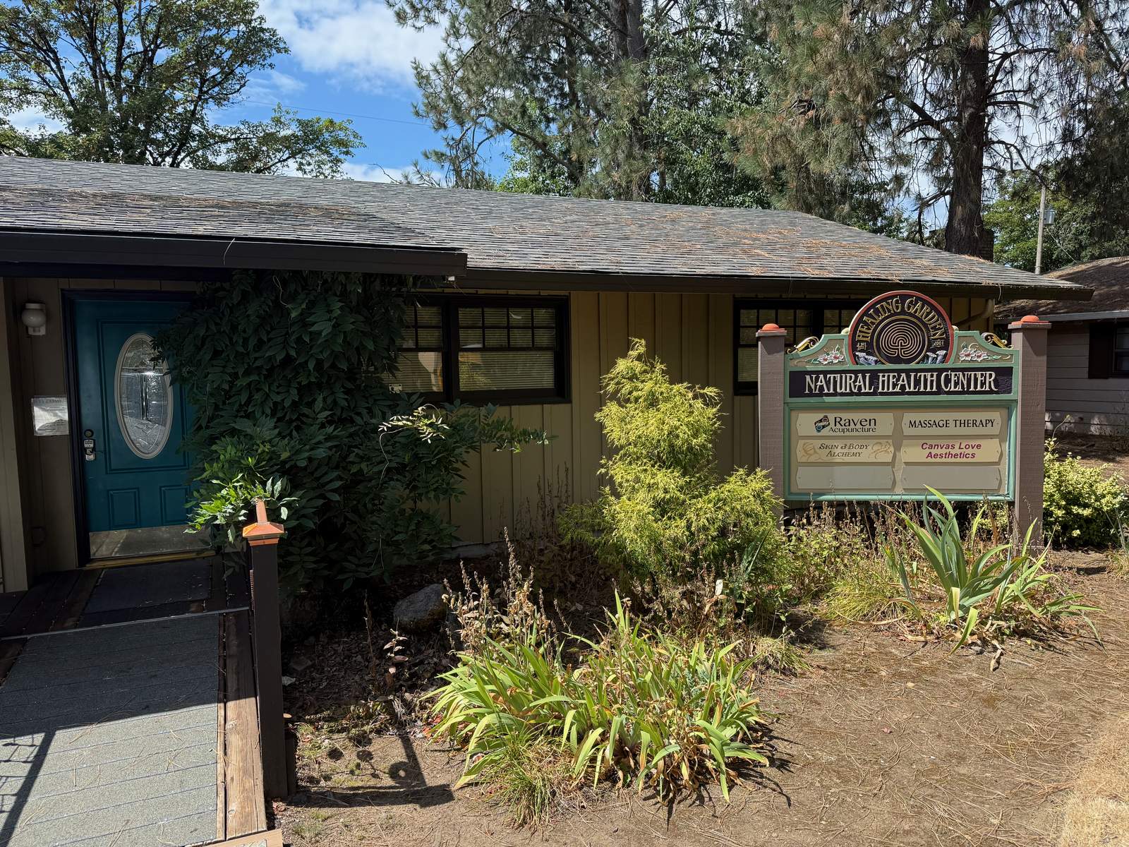A converted-house storefront with a teal front door, ivy climbing a porch post, and a carved wooden sign at the walkway reading "Healing Garden Natural Health Center" above four practice names — Raven Acupuncture, Massage Therapy, Skin & Body Alchemy, and Canvas Love Aesthetics