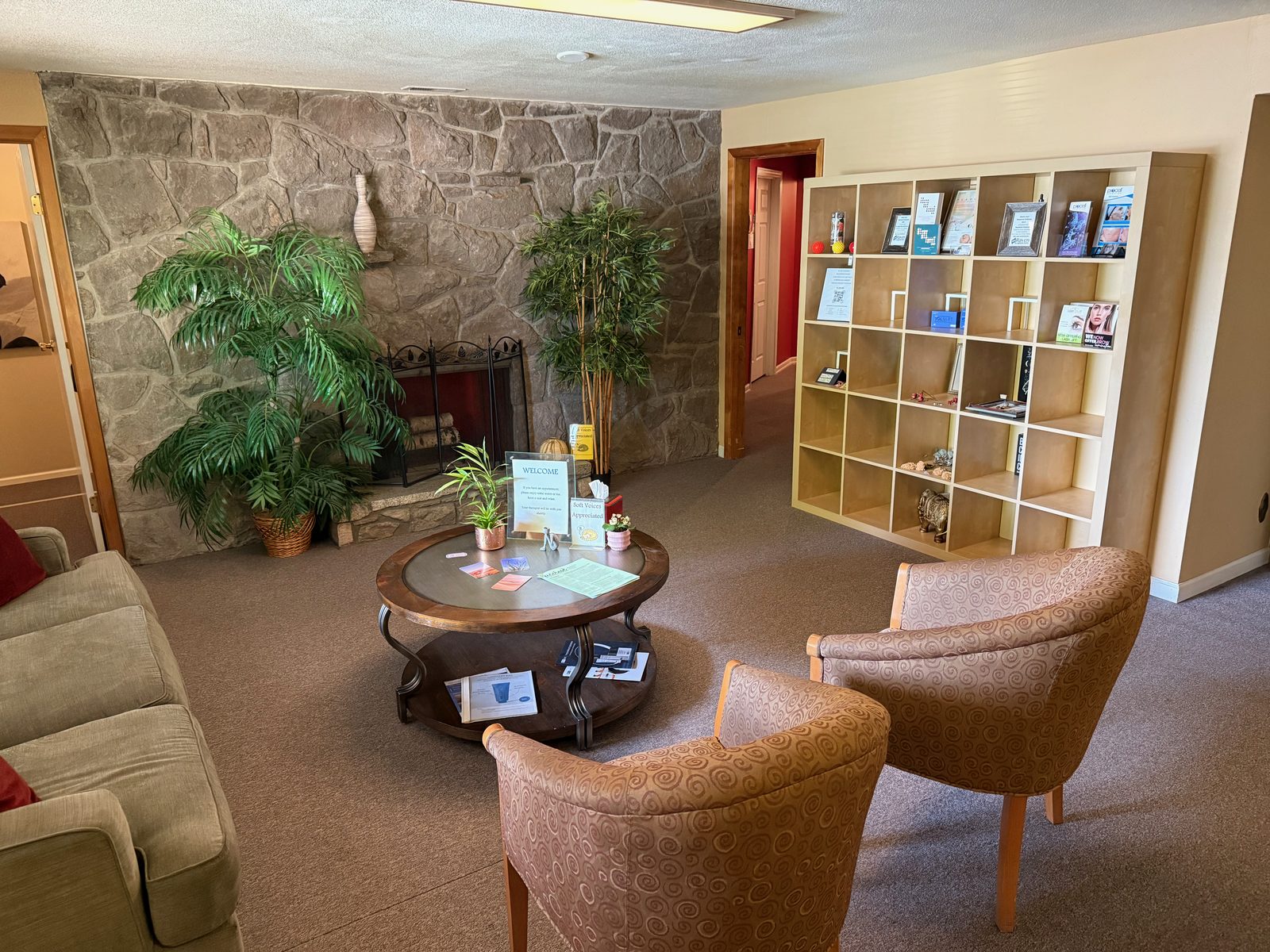 A cozy waiting room with a stacked-stone fireplace wall, two potted palms, a round wooden coffee table with business cards laid out, and upholstered armchairs