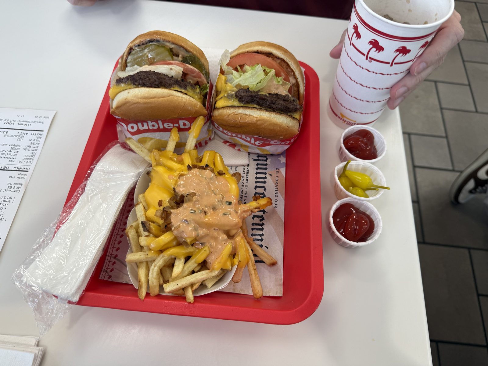 An overhead shot of a red tray holding two halved Double-Double cheeseburgers, a paper boat of Animal Style fries buried in melted cheese and spread, a drink, and three small cups of ketchup and pepperoncini