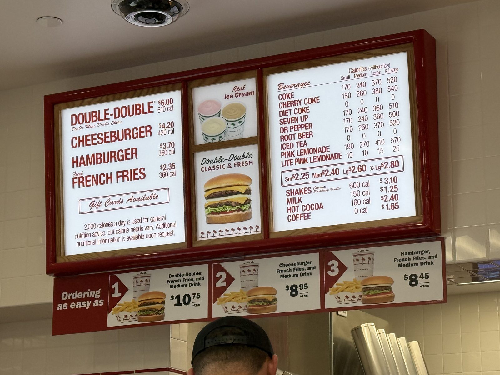 The overhead menu board listing Double-Double $6.00, Cheeseburger $4.20, Hamburger $3.70 and French Fries $2.35, alongside combo pricing and a beverage calorie chart