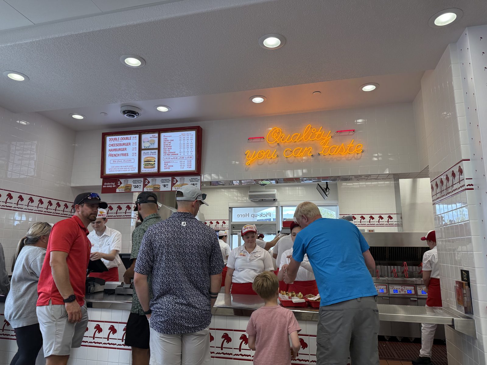 A busy In-N-Out dining room in the late afternoon — a line of customers at the counter beneath an orange neon sign reading "Quality you can taste," with red-and-white palm-tree tile trim along the walls