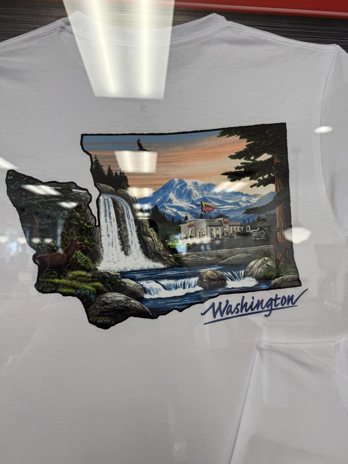A white In-N-Out T-shirt in a display case, printed with an illustration of Mount Rainier, a waterfall and a deer inside the outline of Washington State, with the word Washington in script