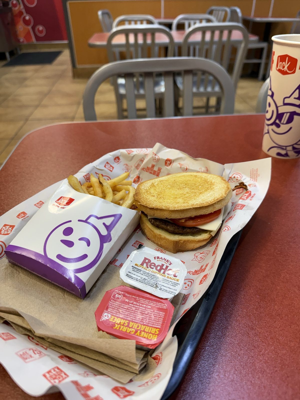 A Sourdough Jack burger and fries on a red tray at Jack in the Box, next to packets of Frank's RedHot buffalo dipping sauce and honey garlic sriracha sauce