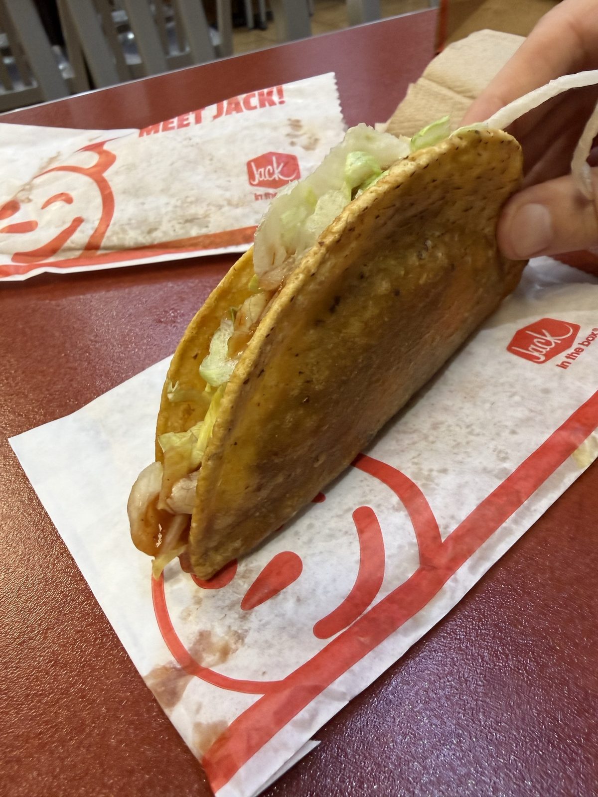 A hand holding up a crunchy Jack in the Box taco with lettuce and shredded cheese, on an unfolded taco wrapper on a red table