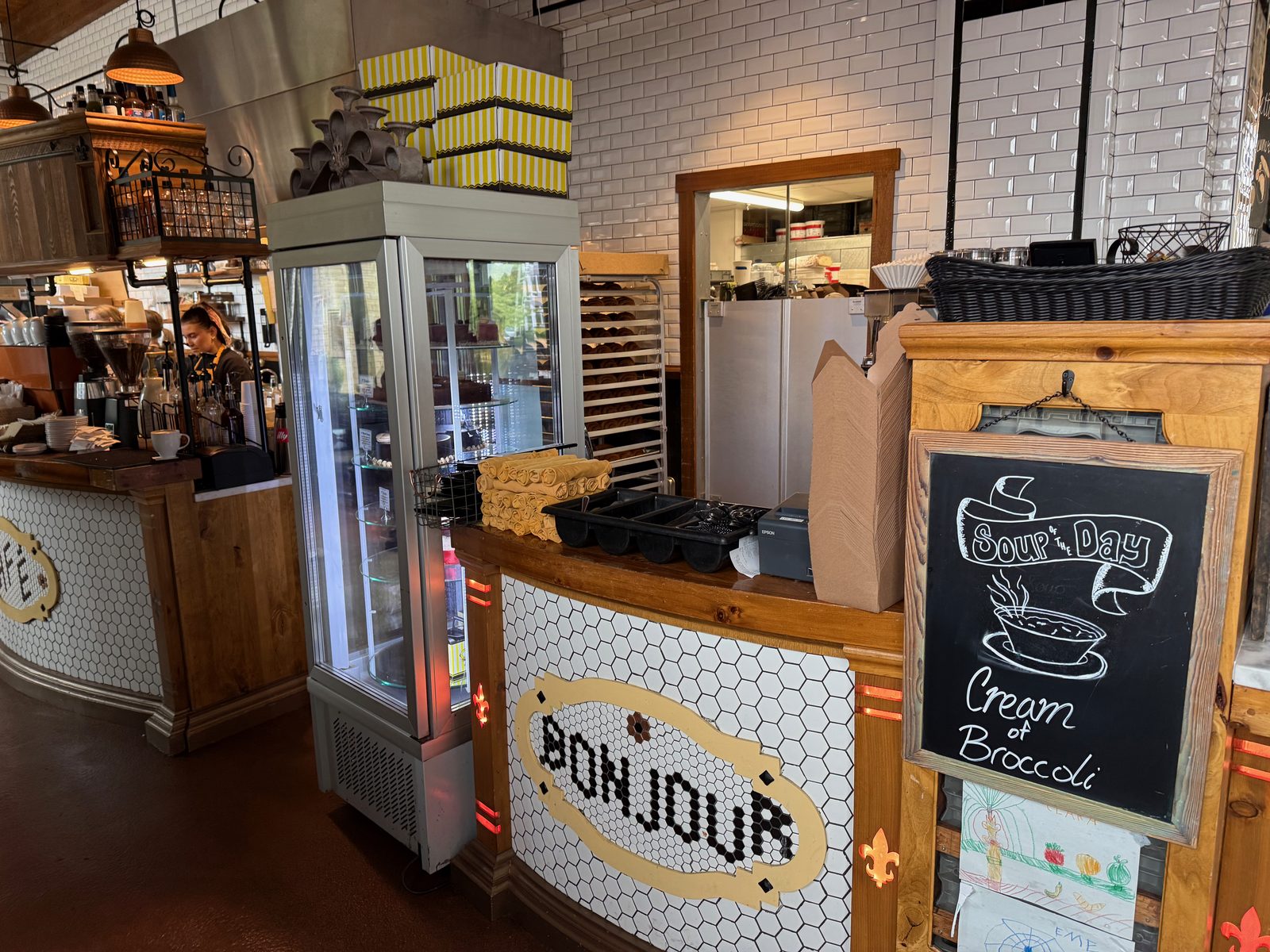 The register area at La Provence with BONJOUR spelled in black tile on white hexagonal mosaic across the counter front, a glass pastry display case, stacked yellow striped bakery boxes above, and a framed chalkboard reading Soup of the Day, Cream of Broccoli