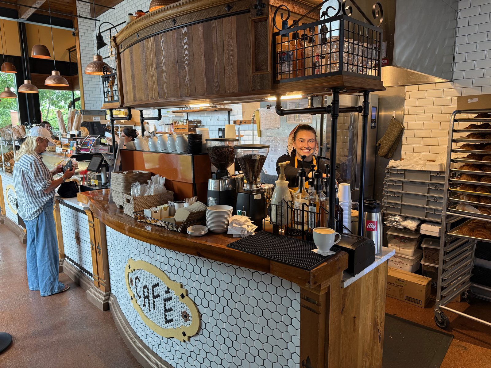 The café counter at La Provence, with CAFÉ spelled out in black tile on white hexagonal mosaic across the front, a barista in an orange apron smiling behind an espresso machine, syrup bottles, stacked cups, a finished latte on the counter and racks of baked croissants to the right