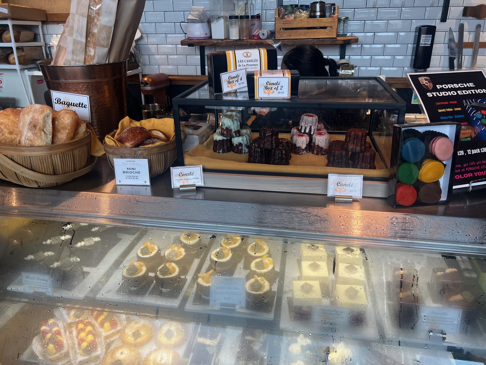 A glass display of canelés at La Provence, dark caramelised fluted pastries arranged in rows, some topped with white icing and berries, with cards reading Canelé 3.75, Canelé Box of 2 at 8.50 and Box of 4 at 17.00, beside baskets of baguettes and mini brioche, and a Porsche Studio Edition macaron box to the right