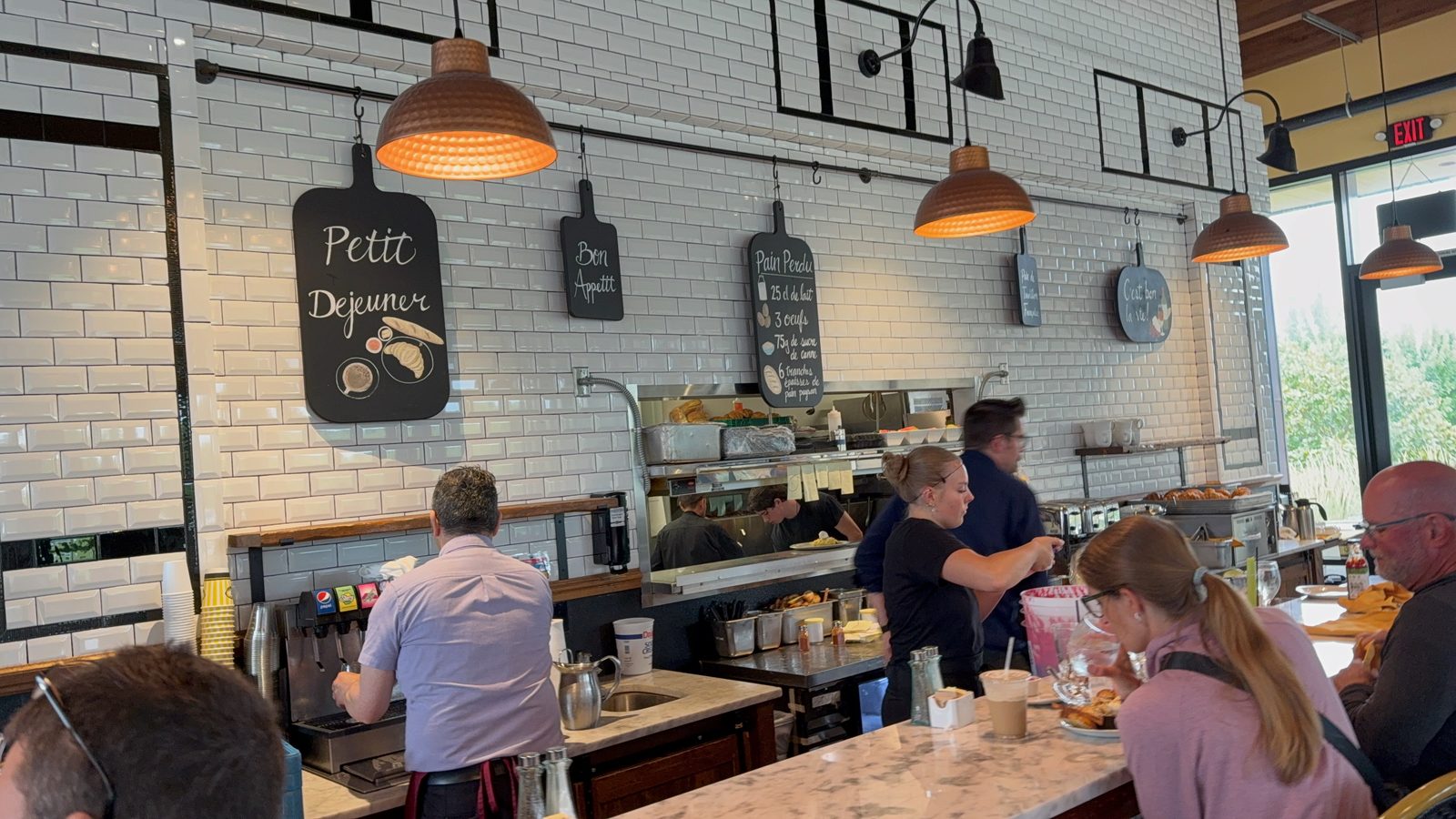 The interior wall at La Provence, white subway tile hung with small black chalkboards reading Petit Déjeuner, Bon Appétit and Pain Perdu, the last with a recipe chalked beneath it in French, copper pendant lamps above, and staff working at the pass below